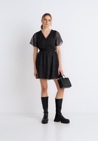 Μαύρο φόρεμα με κοντά, διαφανή μανίκια και ντεκολτέ σε σχήμα V. Το ύφασμα έχει πουα σχέδιο. Συνδυάζεται με μαύρες μπότες μέχρι το γόνατο και μια μικρή τσάντα χειρός.