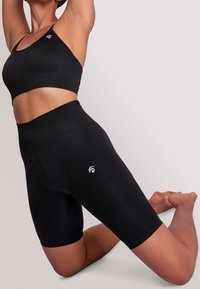 Osoba klečící na jednom koleni, oblečená v černém žebrovaném sportovním podprsence a vysokých cyklistických šortkách na pozadí s jednoduchým světlým pozadím.