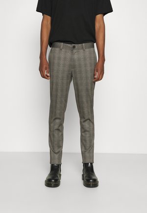 Grijze geruite slim-fit broek met knoop en ritssluiting aan de voorkant, gedragen met zwarte laarzen en een ingestopte zwarte korte mouwen blouse bij een staande persoon.