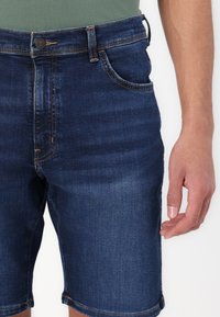 Jeans kratke hlače v temno modri barvi s klasičnim krojem, ki vključujejo sprednje žepke, zadrgo in kontrastne šive.