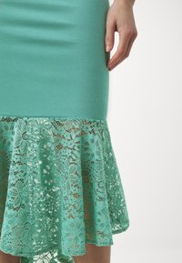 Rochie ajustată de culoare verde mentă cu o margine de dantelă texturată și volane, prezentând modele florale. Țesătură netedă în partea de sus, detalii complicate în partea de jos.