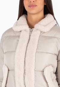 Giacca a vento in beige chiaro con colletto in pelliccia artificiale testurizzato e chiusura frontale con zip. Presenta pannelli cuciti e tasche laterali.
