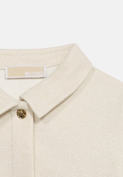 Gulerul texturat bej al cămășii, cu un nasture auriu gravat cu „MK” și o etichetă bej Michael Kors în interiorul gulerului.