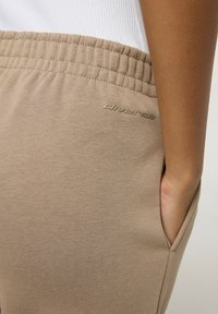 Beżowe spodnie dresowe z elastycznym pasem, boczną kieszenią i subtelnym wytłoczonym logo na udzie. Miękka, teksturowana powierzchnia materiału.