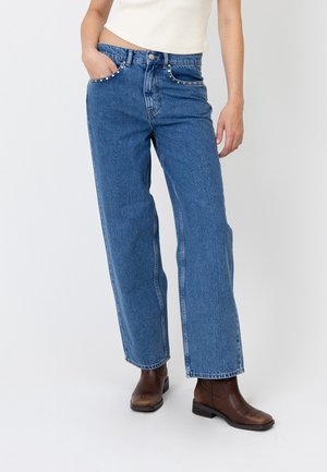 Person iført blå vidde jeans med nittede lommer, en hvid crop top og brune ankelstøvler, står på en ensfarvet hvid baggrund.
