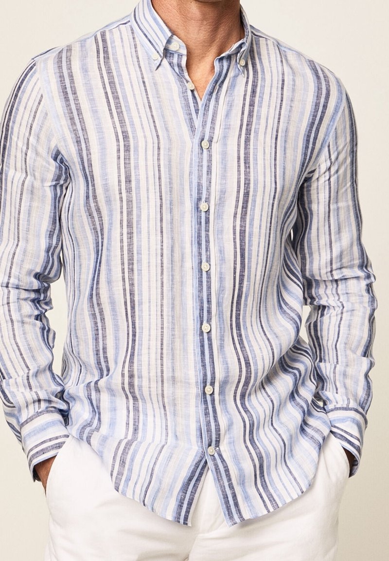 Homme portant une chemise à manches longues boutonnée à rayures verticales bleu, gris et blanc, associée à un pantalon blanc, les mains dans les poches.