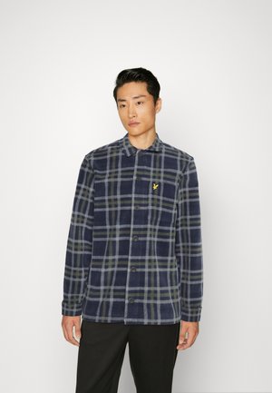 Camicia in flanella a quadri navy con linee verdi e grigie, chiusura a bottoni e un piccolo emblema giallo sul lato sinistro del petto. Maniche lunghe.