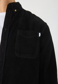 Black corduroy shirt with a button-down collar, featuring a chest pocket and textured ribbing. Minimalist design with subtle branding tag.