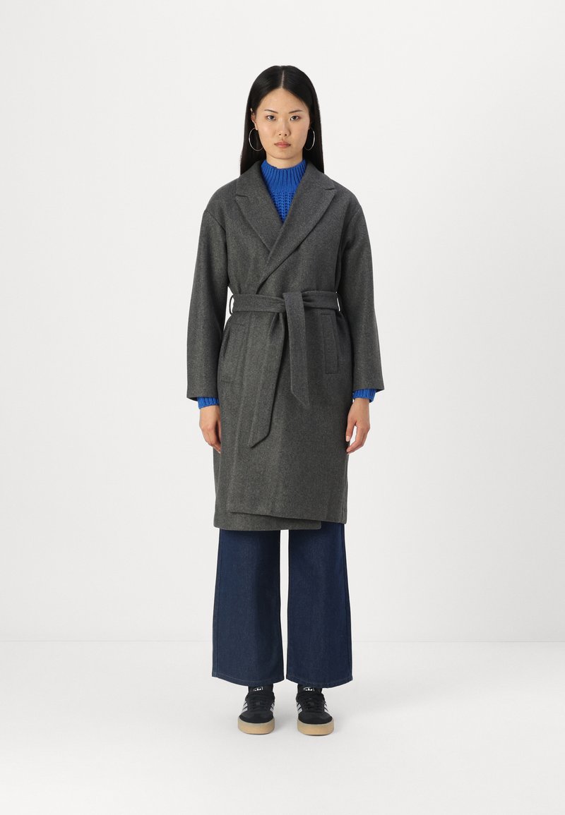 Grå ull omslag frakk med belte, brede slag, og to frontlommer, sammen med en blå ribbestrikket genser og vide denim-bukser.
