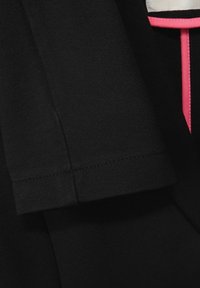 Černá tkanina s hladkou texturou, s růžovým akcentem. Detail rukávu s čistým šitím a strukturovaným tvarem.
