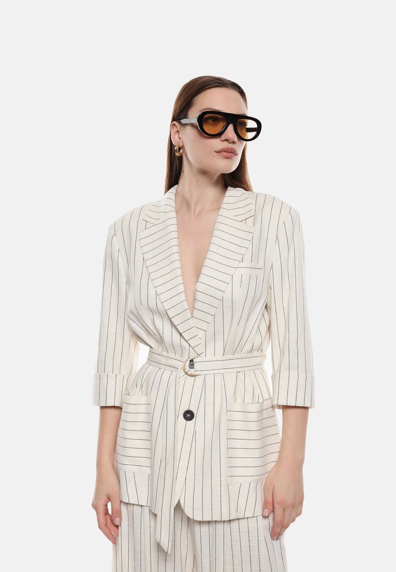 Blazer bianco in lino con righe blu navy, rever a scollo a V, chiusura a un bottone, tasche laterali e una cintura in vita. Indossato con occhiali da sole neri oversize.