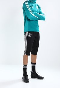 Giacca sportiva a maniche lunghe color teal con tre strisce bianche, pantaloni corti neri con strisce bianche, scarpini neri e calze nere a righe.