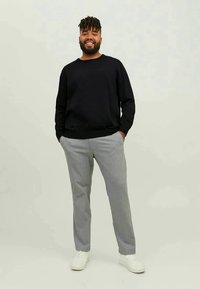 Uomo sorridente in piedi con le mani in tasca, indossa una shirt nera a maniche lunghe, pantaloni grigi a righe e sneakers bianche su uno sfondo neutro.