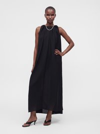 Femme aux cheveux courts portant une robe longue noire sans manches, des sandales noires à talons et un collier fin, debout avec une main sur la hanche.