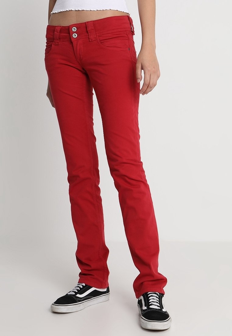 Pantalon rouge skinny à double boutonnière porté avec des baskets noires et blanches par une personne debout sur un fond uni.