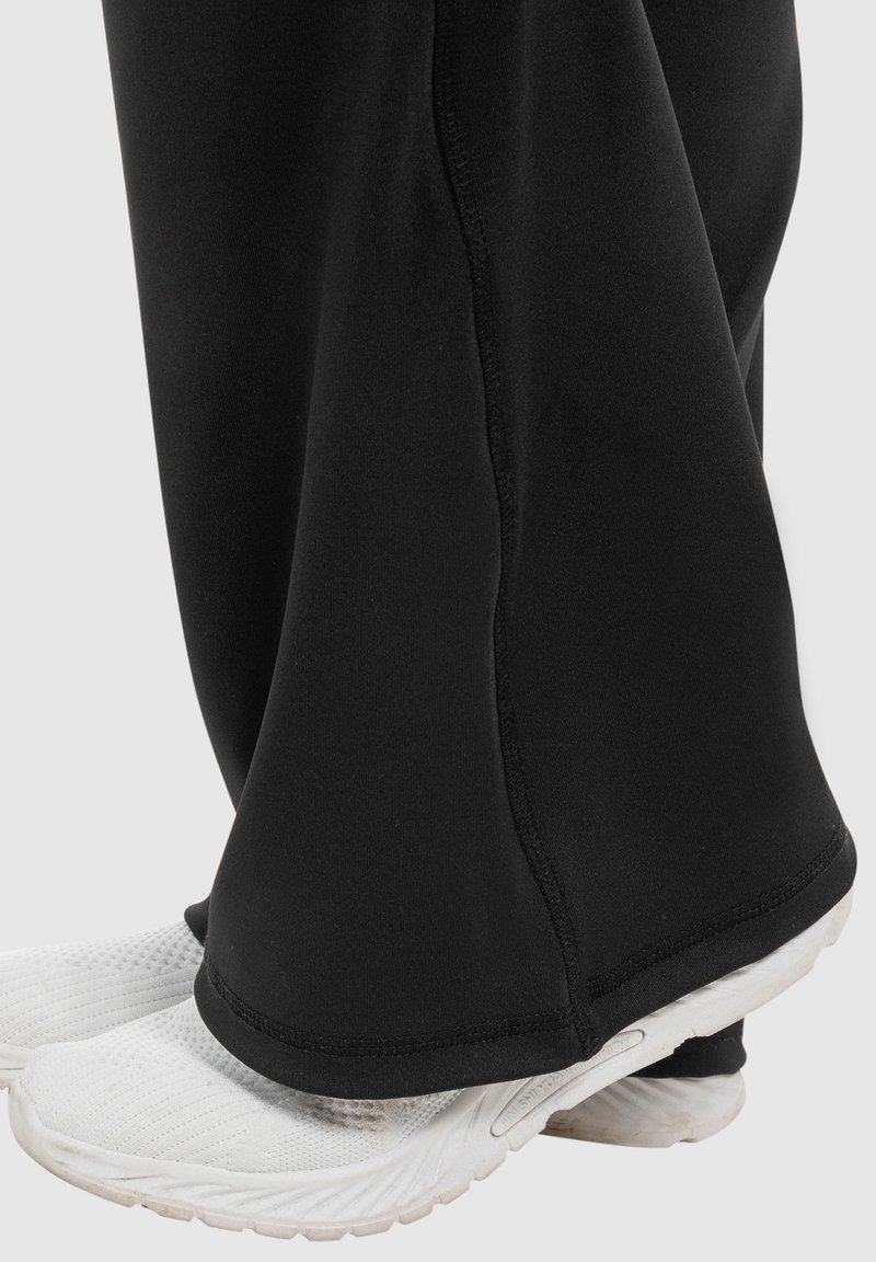 Pantaloni sportivi neri con una vestibilità ampia, realizzati in tessuto morbido e con un taglio dritto, appoggiati su sneakers bianche dalla texture. E.