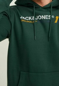 Osoba oblečená v tmavozelené mikině s kapucí, na které je text "JACK & JONES ORIGINALS" a stahovací šňůrky uprostřed, částečně viditelný trup a paže.