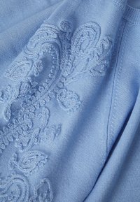 Hele sinine kootud kangas keeruka reljeefse paigutatud paisley-tikkimisega samas toonis sinise niidiga, mis näitab detailset tekstuuri ja õmblusi.