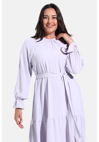Vestido de lila claro con escote redondo, mangas largas y detalles fruncidos. Cuenta con un cinturón de tela en la cintura y un diseño de falda con volantes.