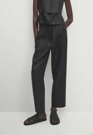 Osoba ubrana w czarny, fakturowany, bezrękawnik, czarne spodnie z wysokim stanem oraz czarne plecione płaskie sandały, stojąca na tle jednolitego tła.