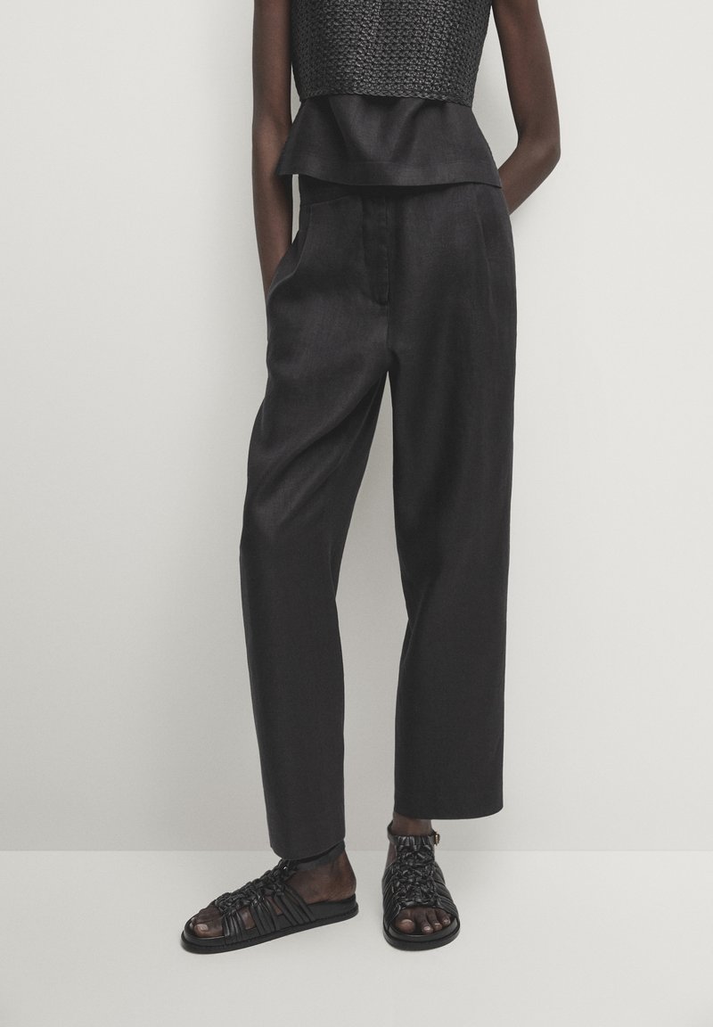 Personne portant un haut sans manches texturé noir, un pantalon noir taille haute et des sandales plates tissées noires, debout contre un fond uni.