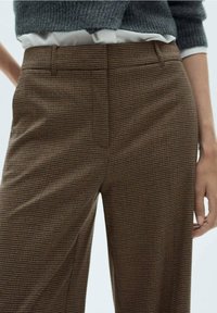 Pantalon à carreaux marron en coupe décontractée, présentant une texture lisse et une taille haute, avec des poches latérales et des détails de couture subtils.