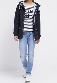 Chaqueta de marine con capucha y forro azul celeste, camiseta de rayas blanca y navy que presenta un gráfico de un gato negro, pantalones vaqueros azules claros y zapatillas blancas.