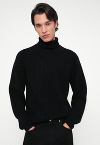 Jeune homme aux cheveux bruns portant un pull à col roulé noir côtelé et un pantalon noir, se tenant avec les mains dans les poches sur fond blanc.