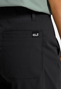 Nahaufnahme der Gesäßtasche einer schwarzen Jack Wolfskin Hose mit Logo-Patch, getragen zu einem hellgrünen Oberteil.