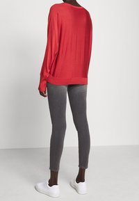 Červený top s dlouhým rukávem z lehké pletené látky, v kombinaci se šedými úzkými kalhotami a bílými teniskami. Jednoduchý design bez výrazných vzorů nebo akcentů.