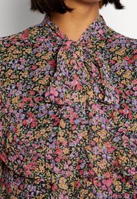 Bloemenblouse in lichtgewicht stof, met een zwarte achtergrond en levendige roze, paarse en gele bloemen. Strikdetail bij de hals.