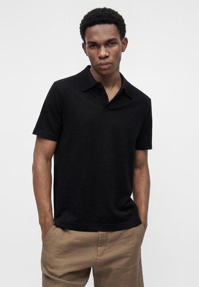 Jeune homme aux cheveux courts et bouclés, portant un polo noir à manches courtes et un pantalon beige, les mains dans les poches, fond neutre.