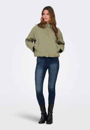 Femme debout, les mains dans les poches, portant une veste zippée vert clair, un jean skinny bleu foncé et des bottines noires sur un fond uni.