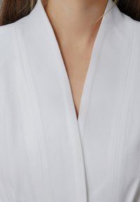 Vêtement blanc texturé avec un décolleté en V profond et un col décontracté. Le tissu semble léger et lisse, mettant en avant un design minimaliste.