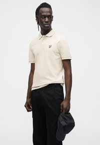 Polo beige con colletto e maniche corte, con logo farfalla giallo. Abbinata a pantaloni neri, con in mano un berretto nero.