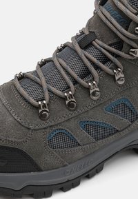 Grau Wanderstiefel aus Wildleder und Mesh-Materialien, mit Metallösen, grauen Schnürsenkeln und blauen Akzenten an den Seiten. Robuste Gummisohle.