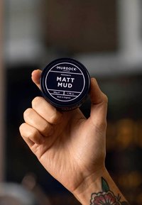 Boîte en métal noir étiquetée "MATT MUD" en texte blanc, tenue dans une main. Fini lisse, avec une zone de prise texturée. Contient 50g.