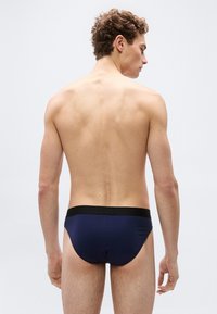 Νέος άνδρας με σγουρά μαλλιά, φαίνεται από πίσω, φοράει σκούρα μπλε εσώρουχα με μαύρη μέση, μπροστά σε ένα απλό ανοιχτό φόντο.