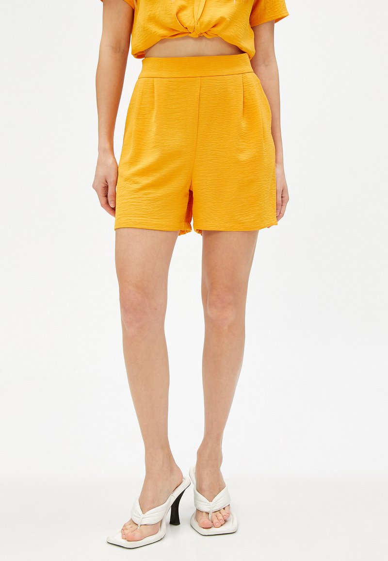 Geel short met een gestructureerde afwerking, elastische tailleband, zijzakken, gecombineerd met witte hoge sandalen. Eenvoudig, schoon ontwerp.