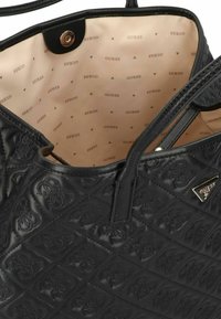 Sac à main synthétique matelassé noir avec un intérieur beige, présentant une doublure à motif et un accent logo métallique.