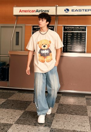 Camiseta oversized de color beige claro con un gráfico de un oso de peluche, combinada con pantalones de mezclilla sueltos de color azul claro y zapatillas blancas de suela gruesa.