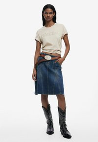 T-shirt in cotone beige con maniche arrotolate, abbinato a una gonna in denim blu fino al ginocchio con cuciture a contrasto e tasche laterali, accentuato da una cintura marrone e stivali cowboy neri.