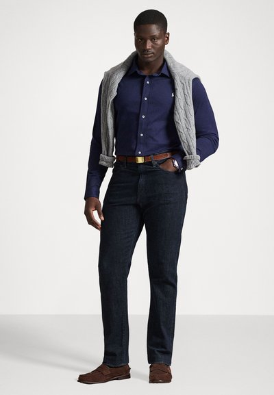 Σκούρο μπλε πουκάμισο με κουμπιά, γκρι πλεκτή ζακέτα ριγμένη στους ώμους, σκούρες τζιν παντελόνες, καφέ παπούτσια και καφέ ζώνη. Ορατό απλό ρολόι.