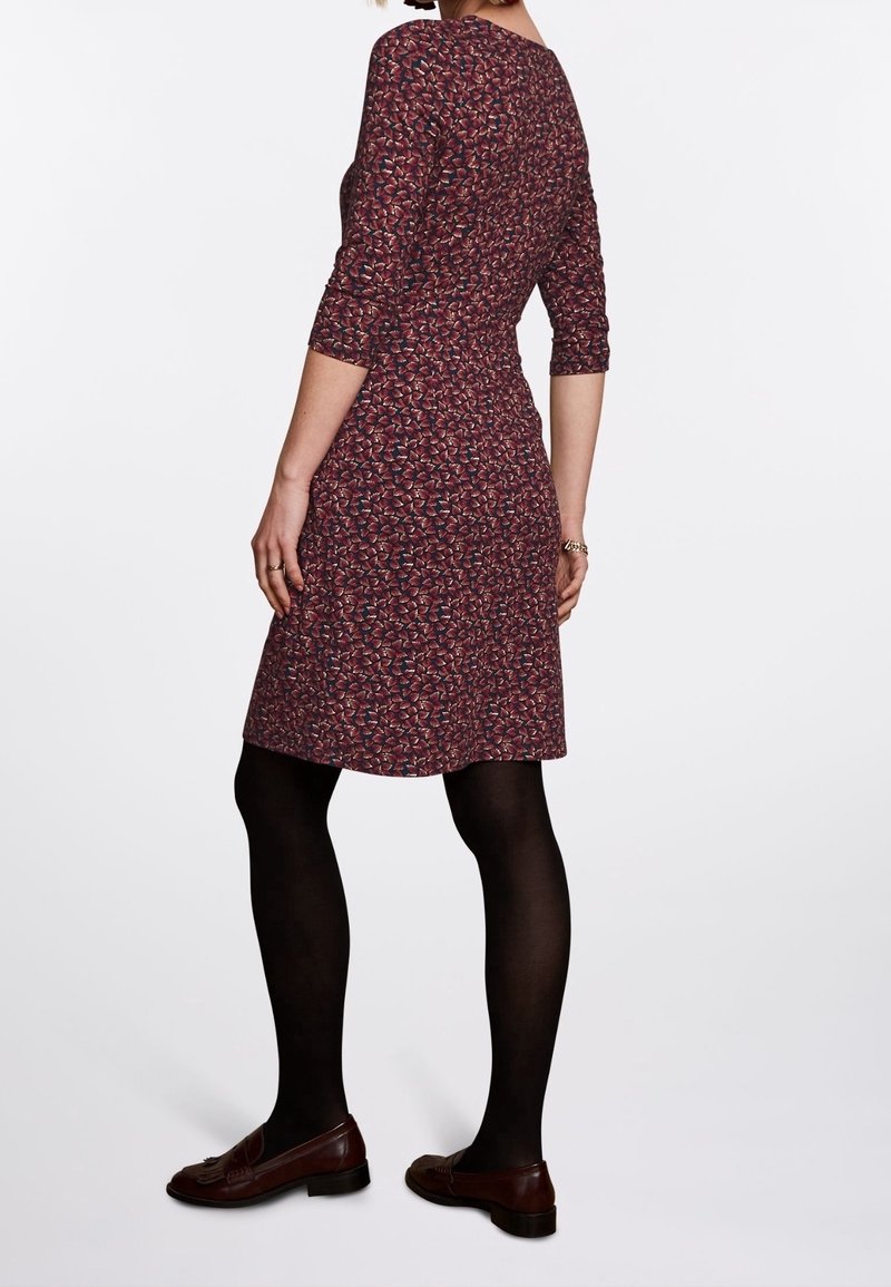 Femme portant une robe fleurie bordeaux mi-longue à manches trois-quarts, des collants noirs et des mocassins marron, debout devant un fond blanc.
