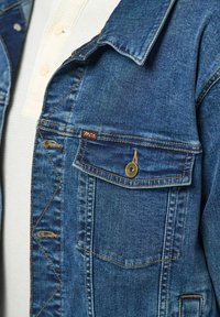 Nahaufnahme einer Tasche aus blauem Denim mit Messingknopf und braunem Etikett, getragen über einem weißen Waffelstrickhemd mit Knöpfen.