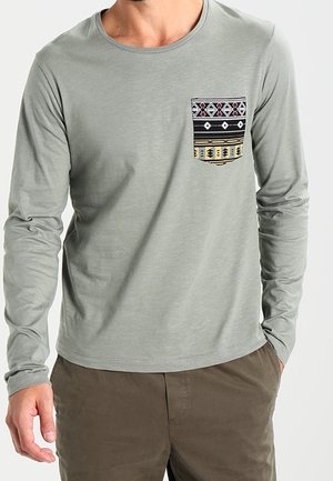 Homme portant une chemise à manches longues gris clair avec une poche poitrine à motifs noirs, jaunes et blancs, et un pantalon marron, les mains dans les poches.