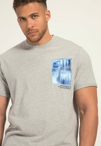 Camiseta de algodón gris con un diseño gráfico azul en el lado izquierdo del pecho, con imágenes y texto sobre "Utopía" y "Otra realidad."
