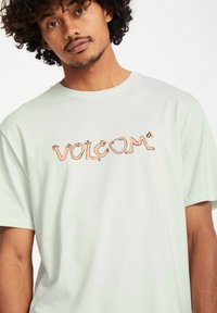 Ljusgrön kortärmad t-shirt med ett tryckt logo "VOLCOM" i orange, med en lekfull handdesign. Mjuk tygstruktur.
