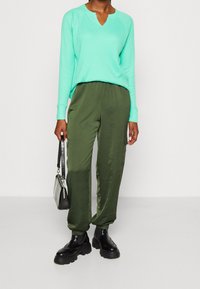 Miętowa bluzka z długim rękawem, z dekoltem w serek i prążkowaną teksturą, zestawiona z oliwkowymi spodniami z elastycznym pasem i czarnymi butami na grubej podeszwie.