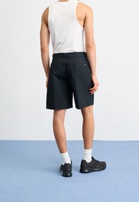 Man som bär svarta shorts, vit linne, vita strumpor och svarta sneakers står på blått golv mot vit vägg, vänd bort.